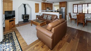 Een woonkamer met een bruine leren bank, een houten salontafel en een televisie. Er is een open haard, een keuken met houten kastjes, en een eettafel met stoelen.