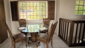 Een eetgedeelte met een ronde glazen tafel, vier houten stoelen en een raam met luiken.