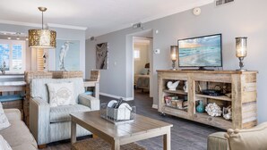 A living room with a TV, wicker furniture, and a wooden cabinet with decorative items. - 🌅 Islander #307 – Sunrise Coffee & Front-Row Beach Action! 🌊🏄‍♂️ (Destin)