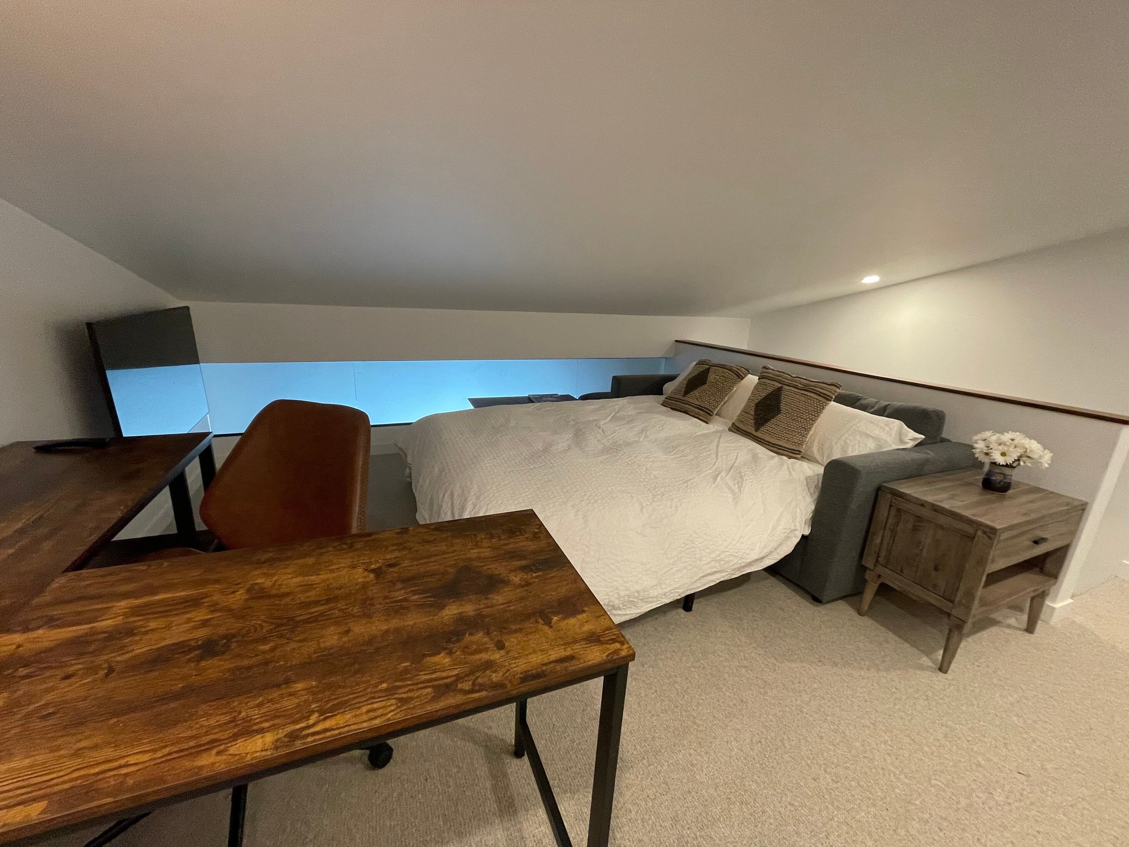 Una habitación de hotel moderna con una cama grande, un escritorio de madera, una mesita de noche pequeña de madera y vistas del cielo a través de una ventana.