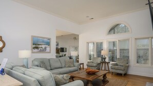 A spacious living room with a large sofa, two armchairs, a coffee table, and a dining table. There are large windows with arched tops and a ceiling fan.