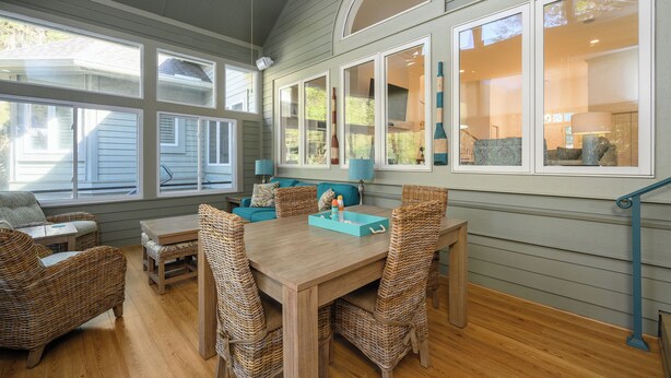 A dining area with a wooden table and wicker chairs, large windows, and a ceiling fan.