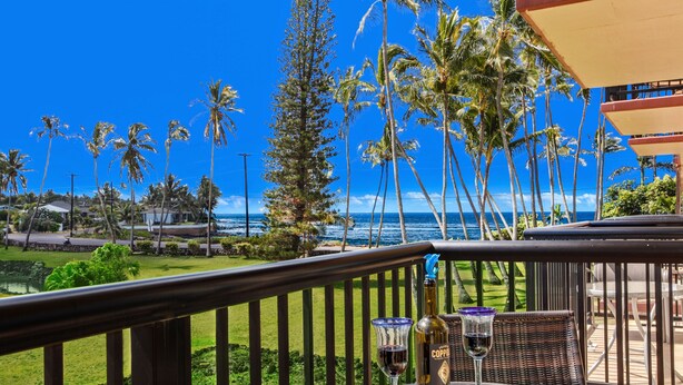 Une table en verre sur laquelle se trouvent une bouteille de Coppa, un verre de vin rouge et une assiette de biscuits, le tout placé sur un balcon donnant sur une plage bordée de palmiers et l’océan.