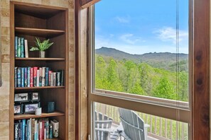 Una habitación con una estantería, un ventanal con vistas a un bosque y montañas, y un balcón con sillas.