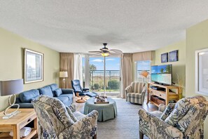A spacious living room with a sofa, chairs, a coffee table, and a television. There is a balcony with a view of the outdoors.