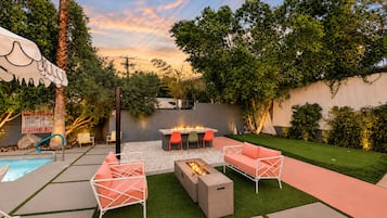 Un patio trasero con piscina, mobiliario exterior, un hogar para la chimenea y un césped bien cuidado.