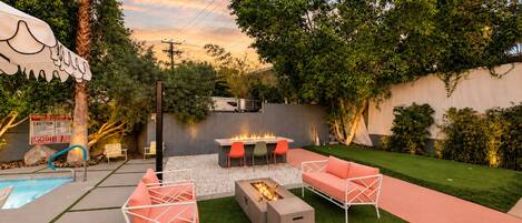 Un patio trasero con piscina, mobiliario exterior, un hogar para la chimenea y un césped bien cuidado.