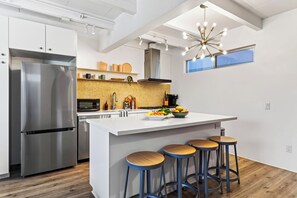 Una cocina moderna con un refrigerador de acero inoxidable, un microondas y un lavamanos. Hay una isla grande con taburetes y un cuenco de fruta.
