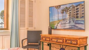 Ett hotellrum med en säng, en fåtölj i rotting, ett träbord med lådor, en tavla som föreställer ett semesterparadis och ett fönster med persienner.