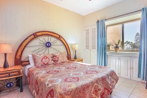 Une chambre avec un grand lit, une table de chevet, une lampe, une fenêtre avec des rideaux et une vue sur des palmiers.