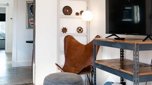 A modern living room with a television, a brown leather chair, a blue ottoman, and a wooden shelf with decorative items. - Unique Limestone Pumphouse + Private Hot Tub (Fredericksburg)
