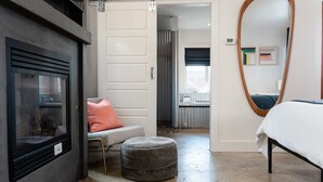 A modern bedroom with a large mirror, a bed, a grey ottoman, and a seating area with a grey sofa and a pink pillow.