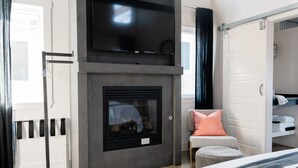 A modern bedroom with a fireplace, a television mounted above it, a closet with a sliding door, and a window with curtains. - Unique Limestone Pumphouse + Private Hot Tub (Fredericksburg)