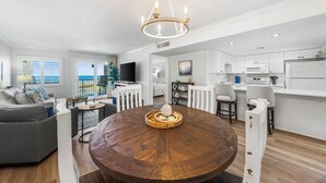 Uma sala de estar moderna com uma mesa de madeira redonda, um sofá cinzento e uma área de cozinha com armários e eletrodomésticos brancos.