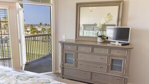 Ein Schlafzimmer mit Bett, Kommode, Fernseher und einem Balkon mit Blick auf Palmen und das Meer.