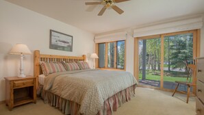 A bedroom with a large bed, two bedside tables, a ceiling fan, and a view of the outdoors through large windows. - JHRL - Teton Pines Townhome #21 (Wilson)