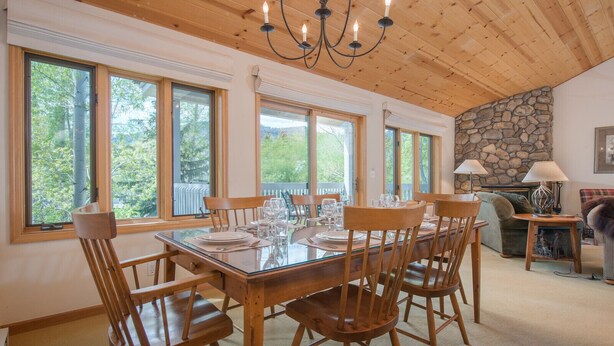 A dining area with a wooden table set for six, surrounded by chairs, large windows, and a stone wall. - JHRL - Teton Pines Townhome #21 (Wilson)