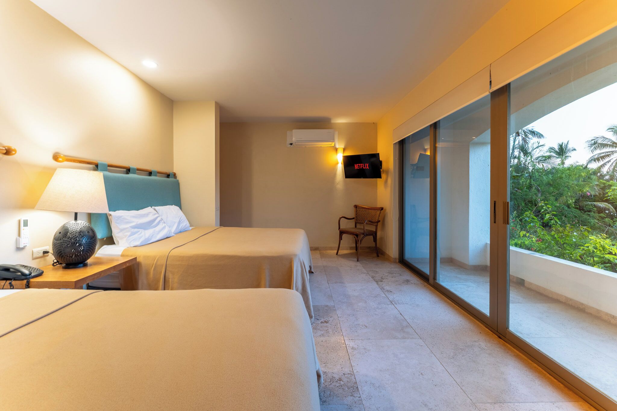 A hotel room with two beds, a bedside table with a lamp, a chair, a wall-mounted TV, and a large window with a view of greenery.