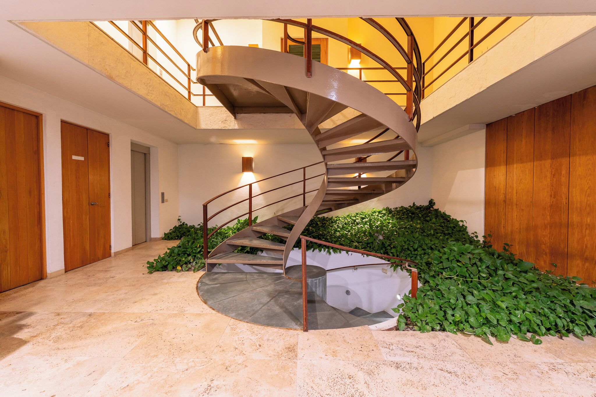 A spiral staircase with metal railings, surrounded by green plants and wooden walls.