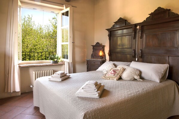 A bedroom with a large bed, a window with a view of trees, a radiator, and a nightstand with a lamp.