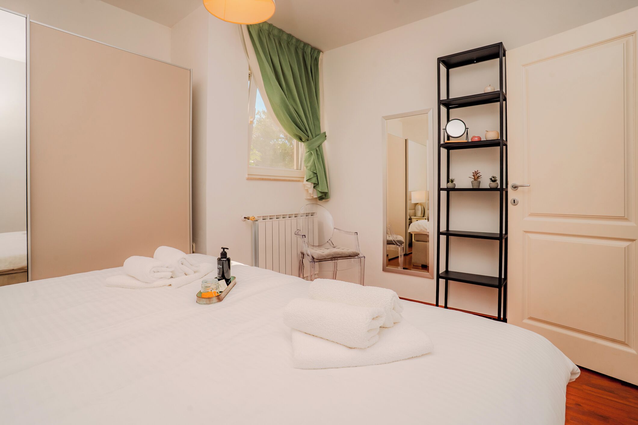 A neatly made bed with white linens, a small table with toiletries, a chair, a radiator, a green curtain, a mirror, and a black shelf with decorative items.