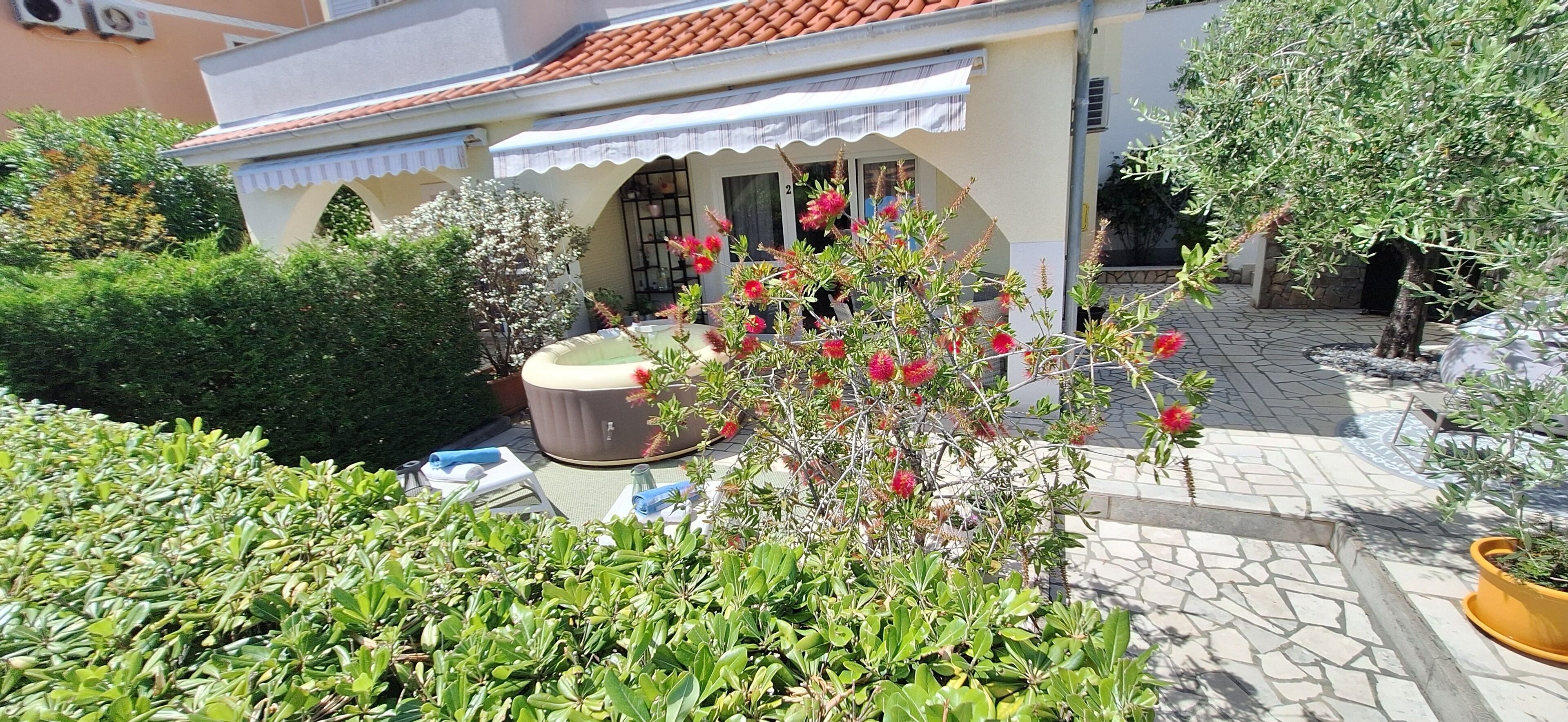 A small building with a tiled roof, a covered porch, and a garden with various plants and a water feature.
