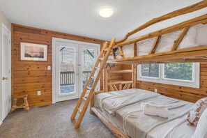 Ein Schlafraum mit Etagenbetten, Holzwänden, einer Stehleiter, einem kleinen Hocker und einer Tür, die auf einen Balkon im Freien führt.