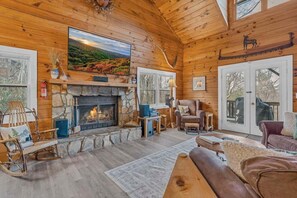A log cabin interior with a stone fireplace, wooden walls, a large window, and a cozy seating area. - Scooby Shack - 10 mins to Main St Blowing Rock, hot tub, pool table (Blowing Rock)
