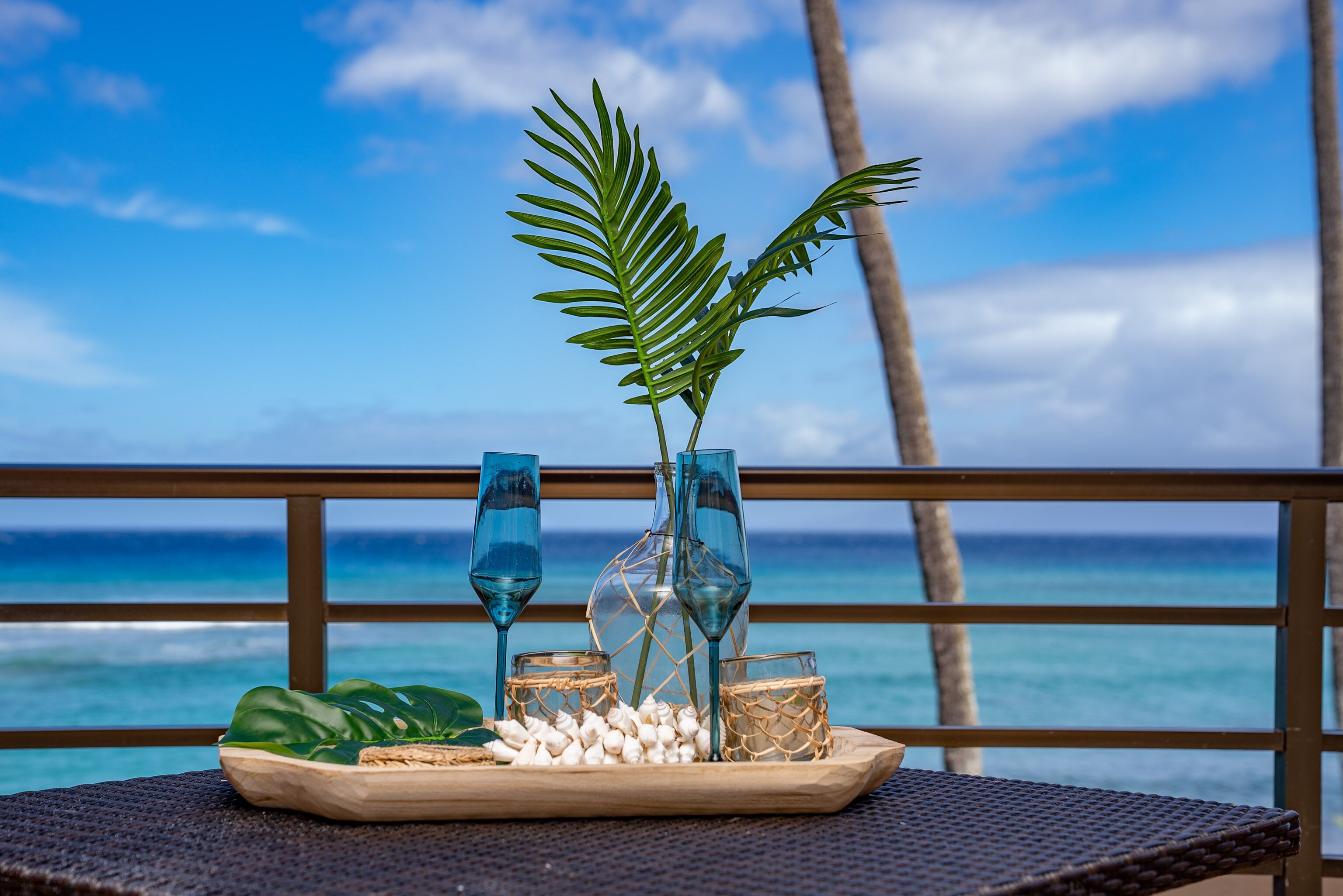 Ett bord med ett bricka fullt av snacks, ett palmblad och två glas vatten, placerat med utsikt över havet och en palm.