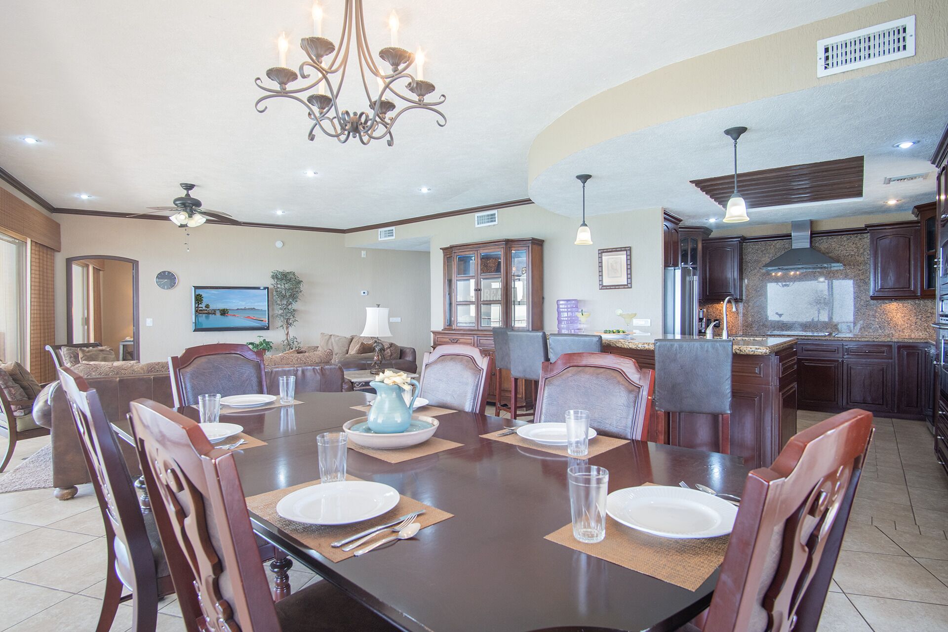 A spacious dining area with a large table set for four, surrounded by comfortable chairs. A chandelier hangs from the ceiling, and the room features a large window and a view of the outdoors.