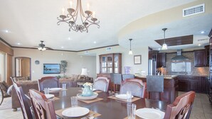 Ruime eetruimte met een grote tafel voor vier personen, omringd door comfortabele stoelen. Een kroonluchter hangt aan het plafond, en de kamer heeft een groot raam en uitzicht op de omgeving.