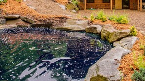 A wooden lodge with a balcony, a stone pond, and a forested backdrop. - 7 Bedroom 5.5 Bath Stunning Luxury Log, Reunions & Weddings Welcomed (Maggie Valley)