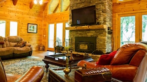 A cozy log cabin interior with a stone fireplace, leather furniture, and large windows. - 7 Bedroom 5.5 Bath Stunning Luxury Log, Reunions & Weddings Welcomed (Maggie Valley)