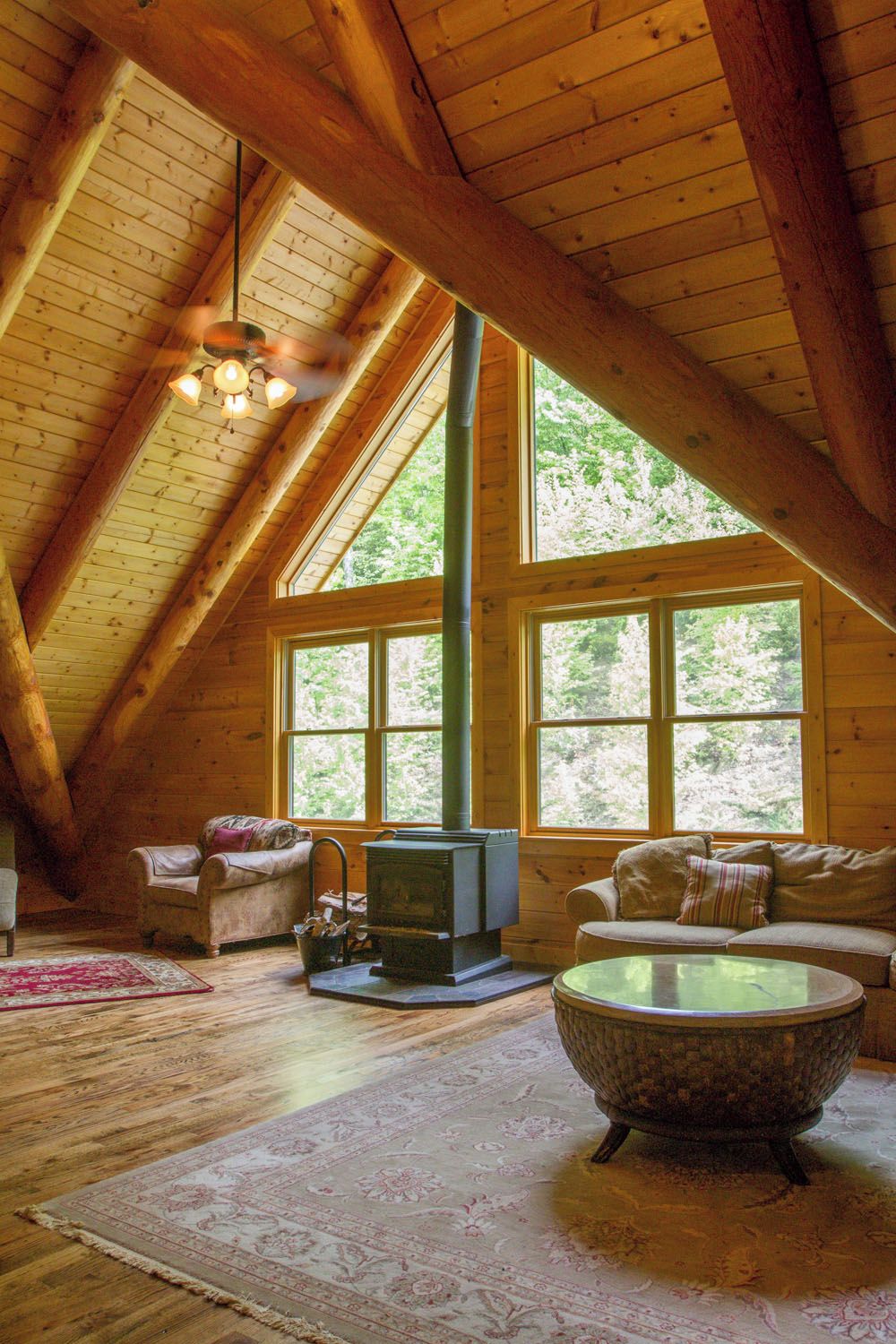 Acogedor dormitorio en ático con vigas de madera, chimenea, dos sofás y un amplio ventanal con vistas a árboles.