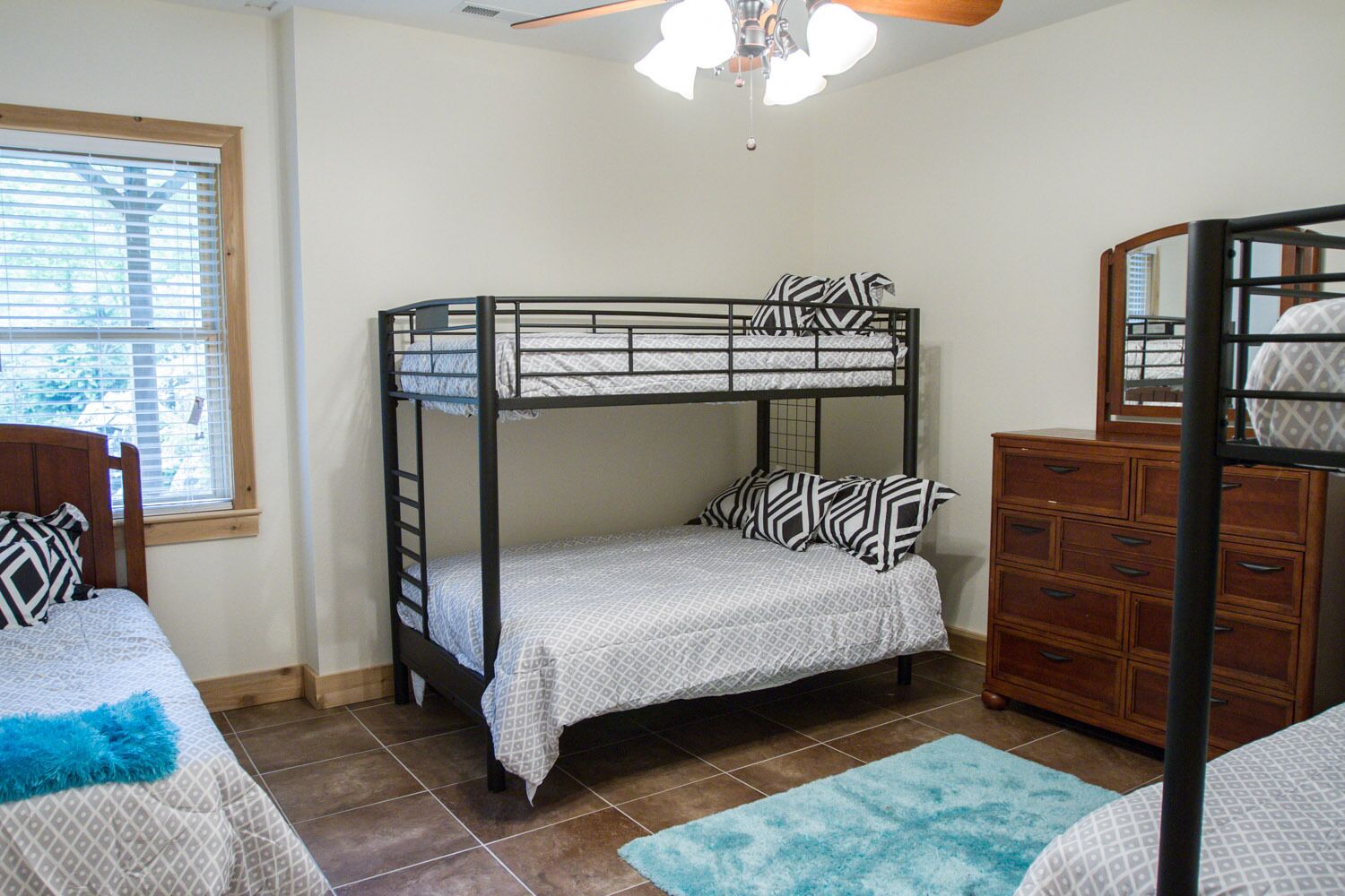 A bunk bed room with a wooden dresser, a window with blinds, and a ceiling fan.