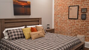A bed with a plaid bedspread and multiple pillows, a wooden headboard, a nightstand with a lamp, and a brick wall with framed items.