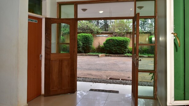 Entrada de un hotel con puerta de madera y paneles de vidrio, que da a un patio con suelo empedrado y vegetación.