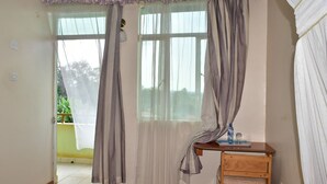 Una habitación con una cama, un escritorio de madera con una silla, una ventana con cortinas y vistas al exterior con vegetación.