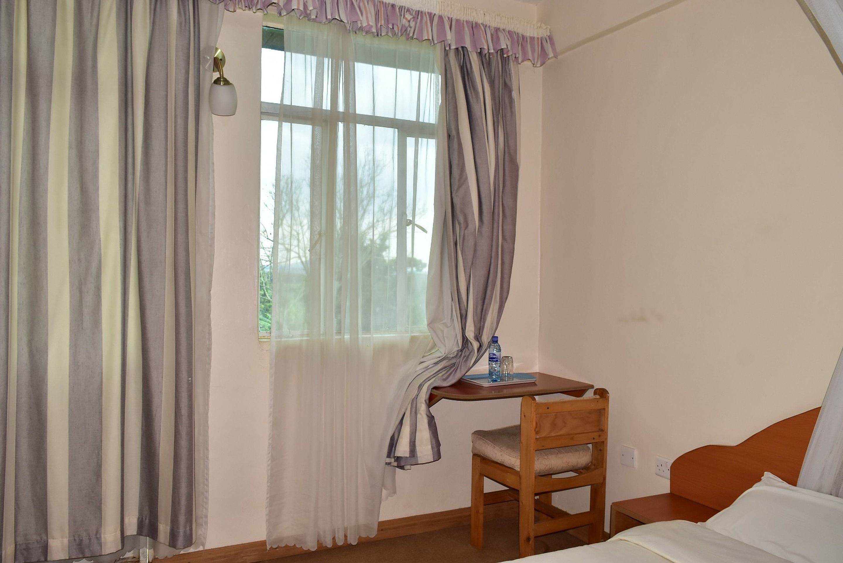 Una habitación pequeña con una cama, una silla de madera, una mesita con una botella de agua y un ventanal con cortinas.