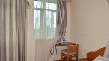 Una habitación pequeña con una cama, una silla de madera, una mesita con una botella de agua y un ventanal con cortinas.