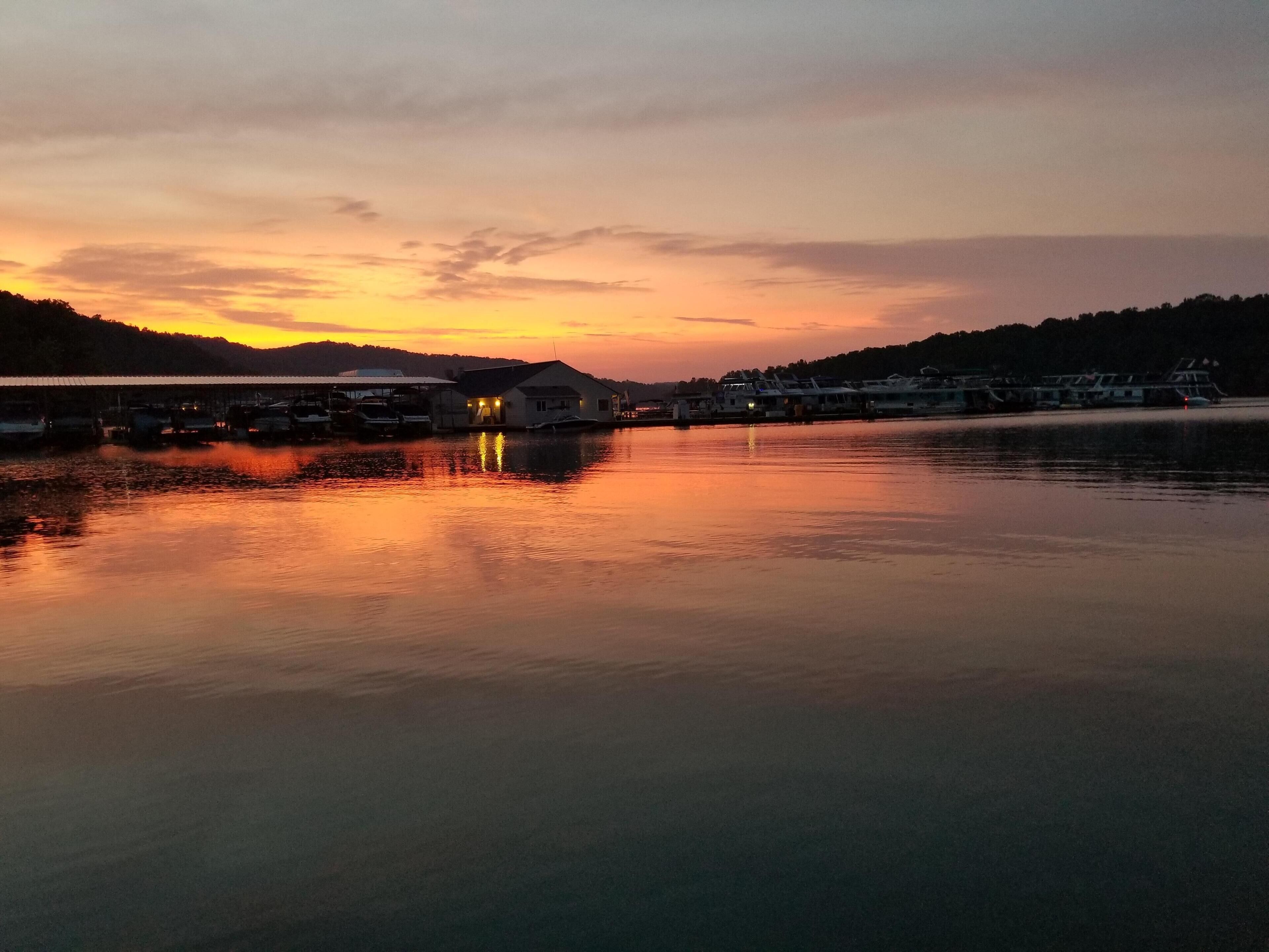 A waterfront area with boats docked, a building with lights on, and a calm sunset.