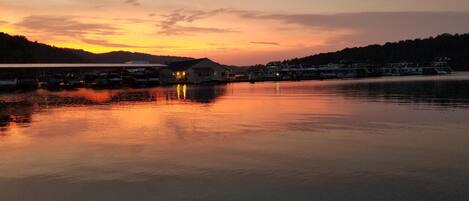 A waterfront area with boats docked, a building with lights on, and a calm sunset.