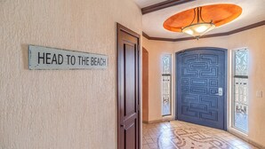 Un couloir avec une pancarte indiquant « DIRECTION LA PLAGE », une porte bleu foncé ornée d’un motif géométrique et un luminaire au-dessus.