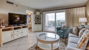 A living room with a TV, a glass coffee table, a sofa, a chair, and a balcony with curtains. - Nice to “Condo” Meet You! Vacation Fun Awaits – Islander 315 (Destin)
