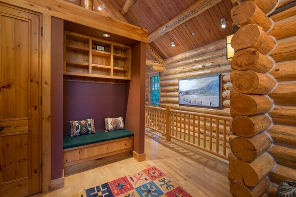 Interior de uma cabana de tronco com um banco de madeira, uma televisão montada na parede e um tapete colorido no chão.