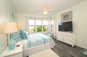A bedroom with a bed, a TV, a ceiling fan, a dresser, and a window with a view of greenery.