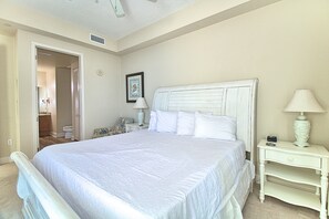 A bedroom with a large bed, a nightstand with a lamp, a framed picture on the wall, and a door leading to another room.