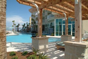 A pool area with lounge chairs and umbrellas, a wooden pergola, and a modern building with balconies.