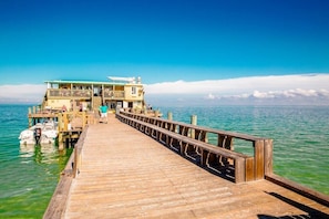 Un muelle de madera que conduce a un edificio de varios pisos con balcones, con vista a una masa de agua tranquila.
