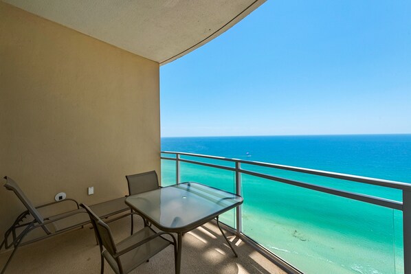 Un balcon avec une table et des chaises en verre, donnant sur une mer turquoise et un ciel dégagé.